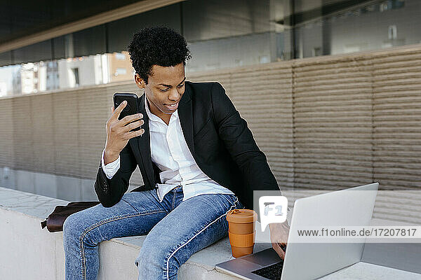 Lächelnder männlicher Unternehmer mit Smartphone und Laptop  der auf einer Stützmauer sitzt