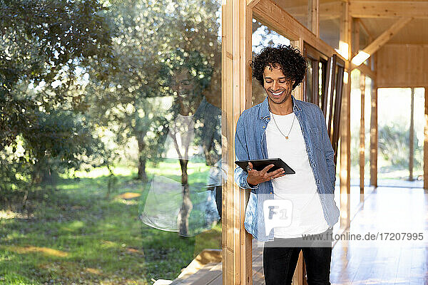 Lächelnder junger Mann  der ein digitales Tablet benutzt und sich an ein Vorgartenfenster lehnt