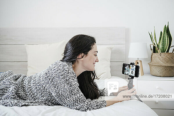Lächelnde Frau im Gespräch über Videokonferenz durch Mobiltelefon  während sie zu Hause auf dem Bett liegt