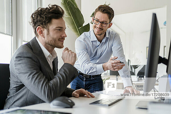 Smiling male colleague discussing with businessman over computer in office