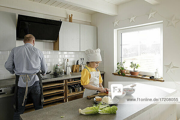 Mädchen mit Kochmütze schneidet Gemüse  während sie mit ihrem Vater zu Hause in der Küche steht