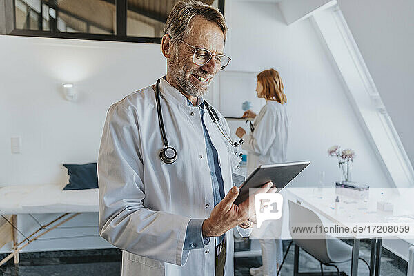 General practitioner using digital tablet while standing with coworker in background at clinic