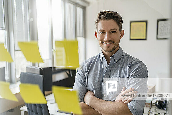 Smiling male entrepreneur with arms seen through glass at office
