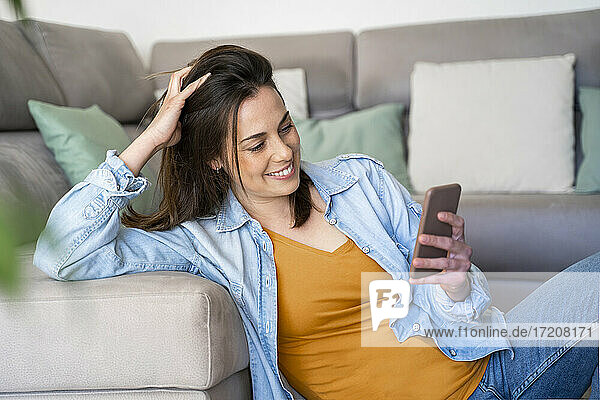 Lächelnde junge Frau  die ein Mobiltelefon benutzt  während sie auf dem Sofa im Wohnzimmer sitzt