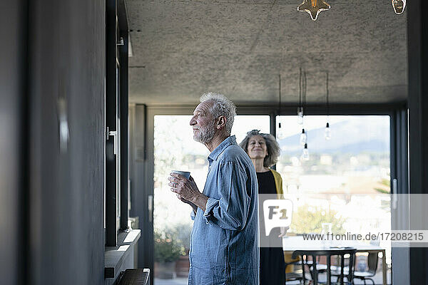 Man holding coffee cup while standing by window with woman in background at home