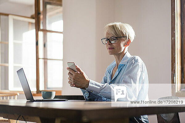 Lächelnde Unternehmerin mit Laptop  die ein Mobiltelefon benutzt  während sie am Schreibtisch im Büro zu Hause sitzt