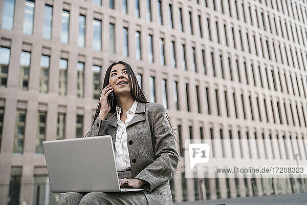 Nachdenkliche Geschäftsfrau  die ein Smartphone benutzt  während sie einen Laptop vor einem Gebäude in der Stadt benutzt