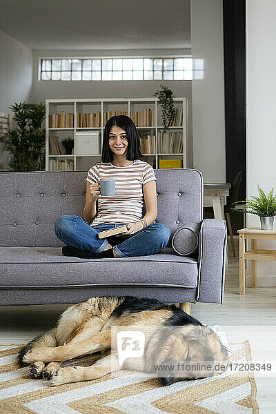 Smiling young woman with coffee cup sitting on sofa while dog sleeping on carpet