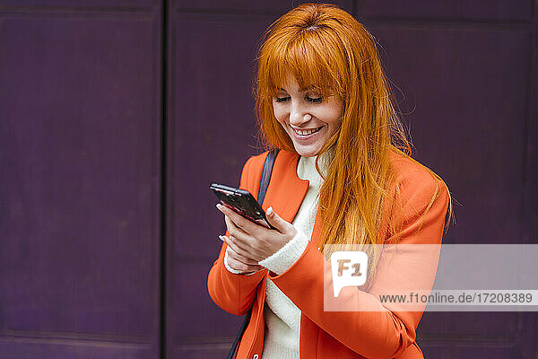 Lächelnde Frau in Jacke mit Mobiltelefon an der Wand