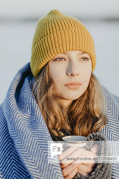 Porträt eines hübschen Mädchens im Teenageralter mit Strickmütze  das sich mit einer Wolldecke und einer Tasse heißem Tee wärmt