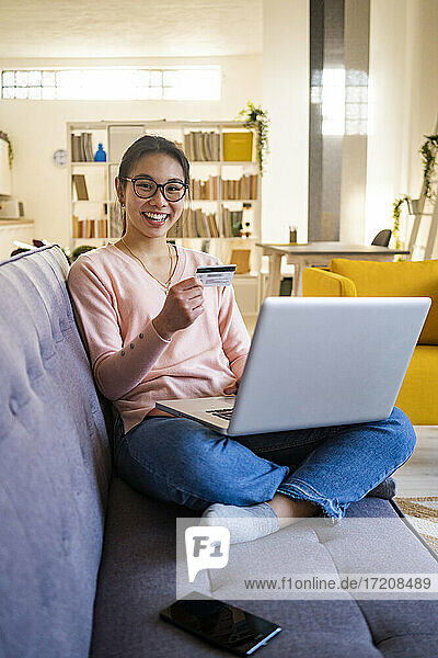Fröhliche Frau  die eine Kreditkarte in der Hand hält  während sie zu Hause am Laptop sitzt