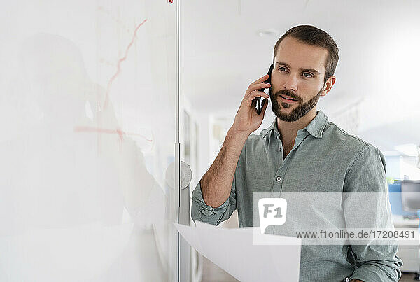 Geschäftsmann  der mit einem Mobiltelefon spricht  während er im Büro vor einem Whiteboard steht