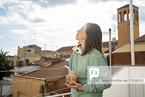 Frau mit geschlossenen Augen  die einen Fruchtsmoothie hält  während sie auf einem Balkon steht
