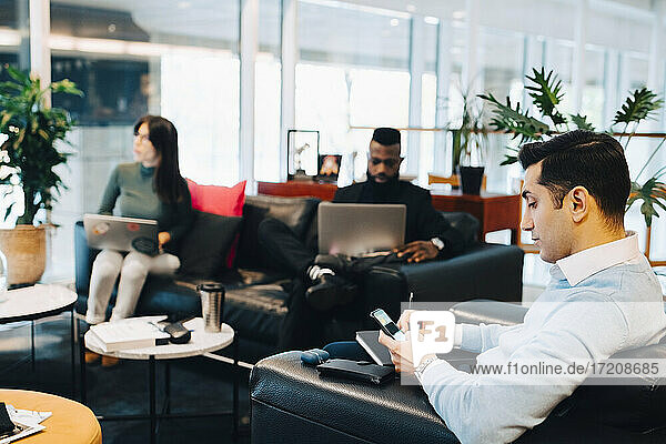 Männlicher Unternehmer bei der Nutzung eines Smartphones während der Arbeit mit Kollegen im Büro