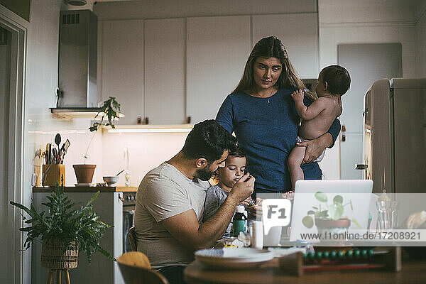 Vater füttert Sohn  während Mutter mit Baby-Mädchen in der Küche steht