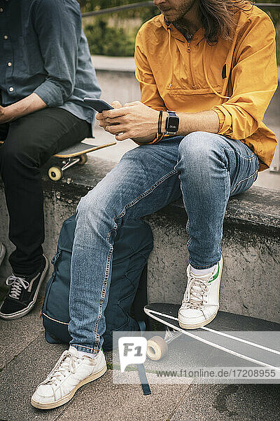 Niedriger Ausschnitt eines Mannes  der ein Smartphone benutzt und neben einem männlichen Freund auf einer Stützmauer sitzt