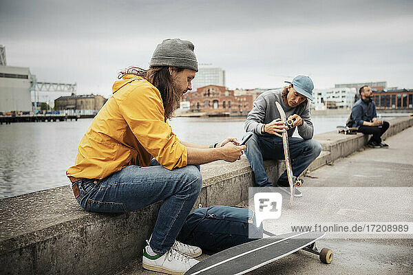Male friends with smart phone and skateboard sitting on retaining wall