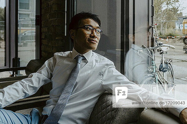 Männlicher Unternehmer schaut durch das Fenster  während er auf dem Sofa im Büro sitzt