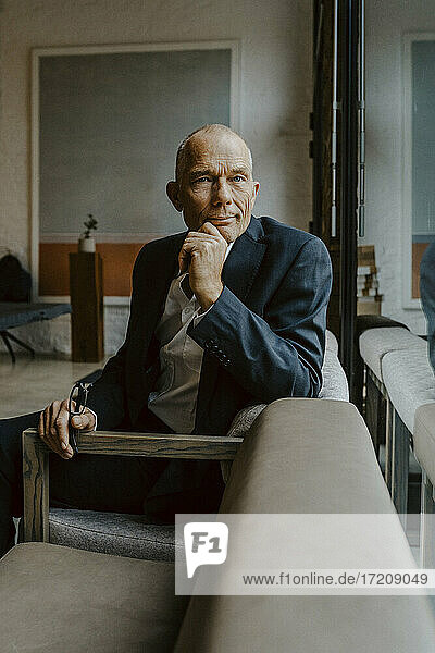 Porträt eines männlichen Unternehmers mit Hand am Kinn  der über einem Sofa im Büro sitzt