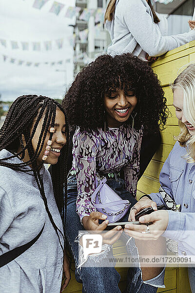 Lächelnde Freundinnen  die ein Smartphone benutzen  während sie auf einer Kiste im Freien sitzen
