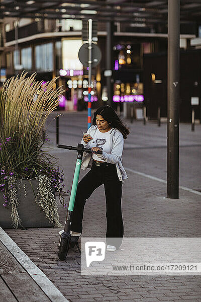 Teenage girl using smart phone while standing with electric push scooter on footpath during sunset