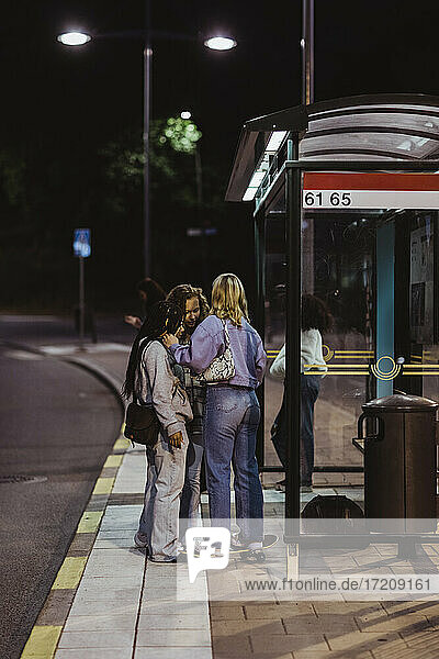 Weibliche Freunde reden  während sie nachts an der Bushaltestelle stehen