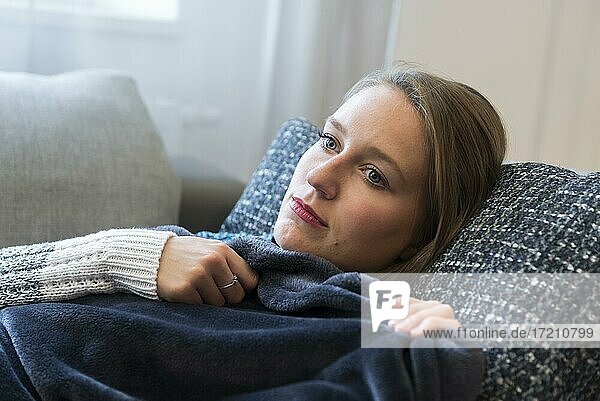 A young woman lies cuddled up in a blanket on a sofa in Dorfen  Bavaria  Germany  Europe