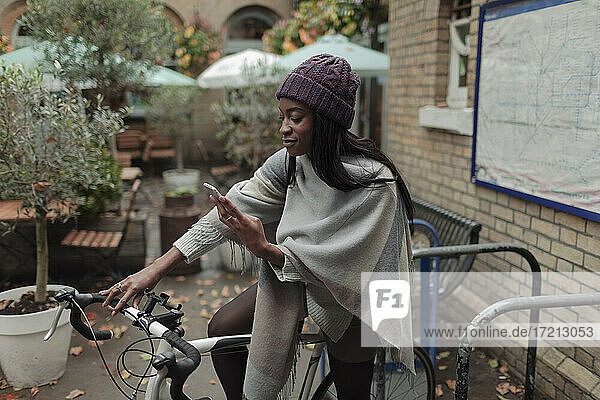 Junge Frau mit Smartphone auf dem Fahrrad vor einem Straßencafé