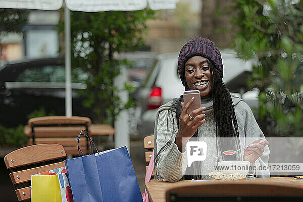 Happy young woman with smart phone eating soup at sidewalk cafe