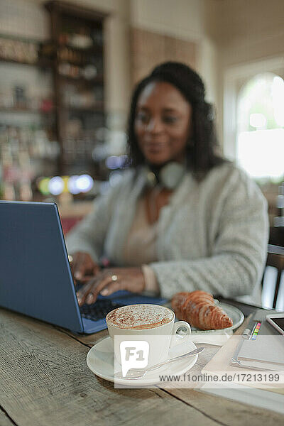 Woman working at laptop next to cappuccino and croissant in cafe