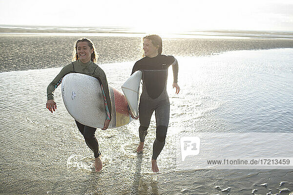 Happy young female surfers running with surfboards in sunny ocean surf