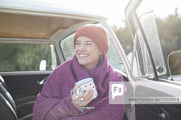 Glückliche junge Frau mit Tee in Decke eingewickelt bei sonnigen van