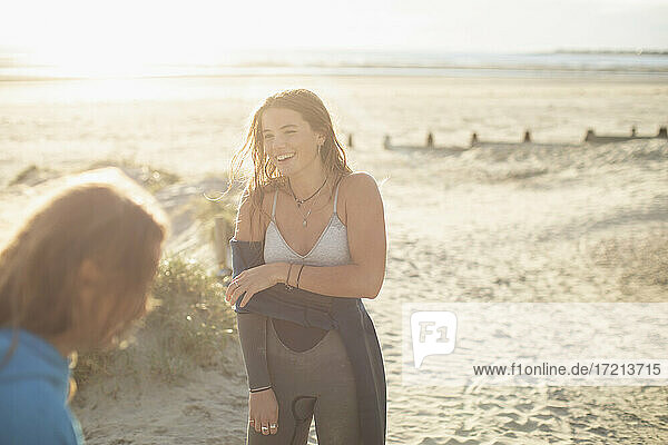 Glückliche junge weibliche Surferin beim Ausziehen des Neoprenanzugs am sonnigen Strand