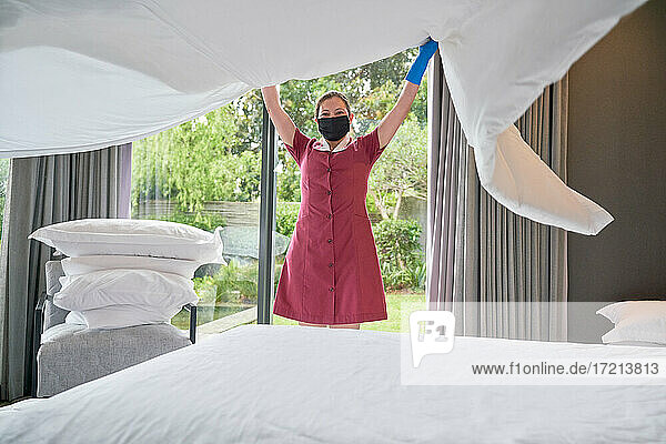 Weibliches Hotelzimmermädchen in Gesichtsmaske macht Bett im Hotelzimmer