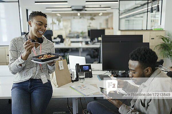 Happy business people eating takeout lunch at desk in open plan office