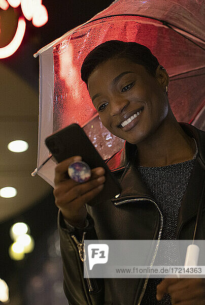 Glückliche junge Frau mit Smartphone unter Regenschirm in der Nacht