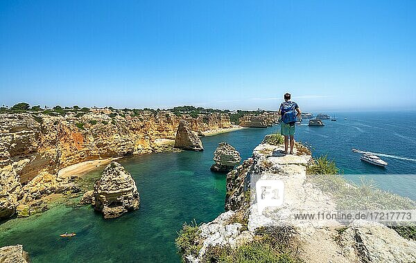 Young man standing on rocks on cliff  view on cliff of sandstone rocks  rock formations in turquoise sea  sandy beach  Praia da Marinha  Algarve  Lagos  Portugal  Europe