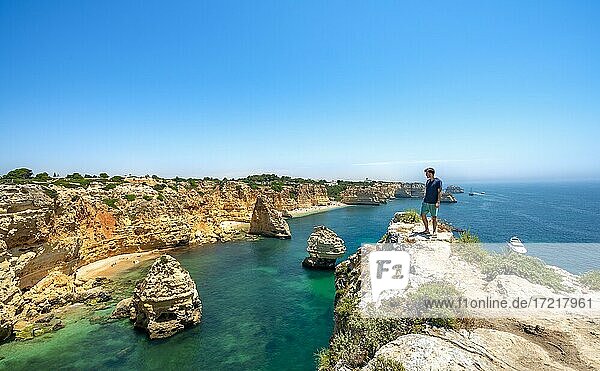 Young man standing on rocks on cliff  view on cliff of sandstone rocks  rock formations in turquoise sea  sandy beach  Praia da Marinha  Algarve  Lagos  Portugal  Europe