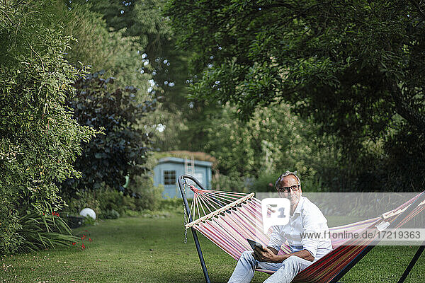 Nachdenklicher Mann mit digitalem Tablet in einer Hängematte im Garten sitzend