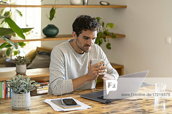Konzentrierter Freiberufler  der einen Kaffee trinkt  während er auf seinen Laptop im Heimbüro schaut