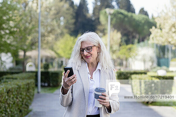 Smiling woman using mobile phone while holding reusable cup at park