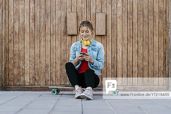 Lächelnde Frau mit Kopfhörern  die ein Smartphone benutzt  während sie auf einem Longboard an einer Holzwand sitzt