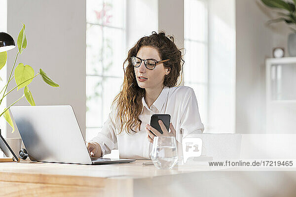 Berufstätige Frau mit Brille arbeitet am Laptop und hält ein Smartphone am Schreibtisch