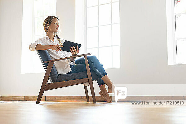 Thoughtful woman looking away while holding digital tablet on chair at window