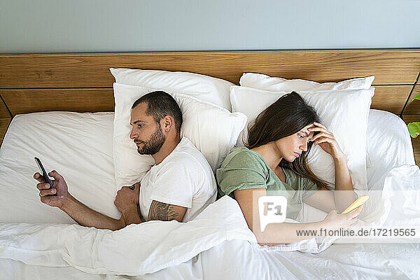 Junges Paar benutzt Smartphones  während es Rücken an Rücken zu Hause im Bett liegt