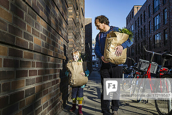 Man with daughter holding paper bag of vegetables while walking on alley