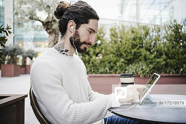 Männlicher Hipster mit Einwegbecher  der während eines Videogesprächs in einem Straßencafé ein digitales Tablet berührt