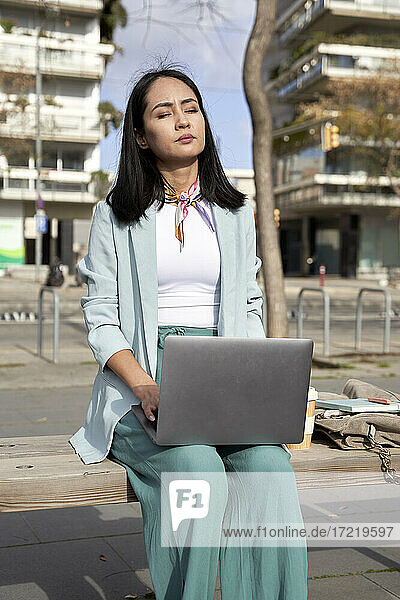 Young businesswoman with eyes closed sitting with laptop on bench