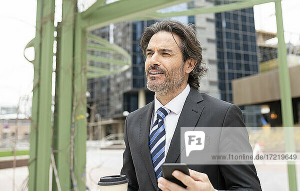 Lächelnder männlicher Berufstätiger  der mit Kaffeetasse und Mobiltelefon in der Hand wegschaut