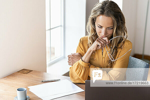 Female professional holding eyeglasses while looking at laptop on desk